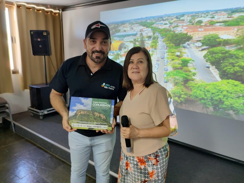 César Siboldi (com barba curta, vestido de calça jeans, camiseta polo preta, com logo da prefeitura de Águas de São Pedro e boné preto) recebe, em nome da Região Turística Serra do Itaqueri, um exemplar do Livro Geoparque Corumbataí das mãos da Profa. Mariselma Zaine (cabelos loiros na altura do ombro, vestida de saia com estampa de flores e camisa de linho beje). Foto de André Kolya.